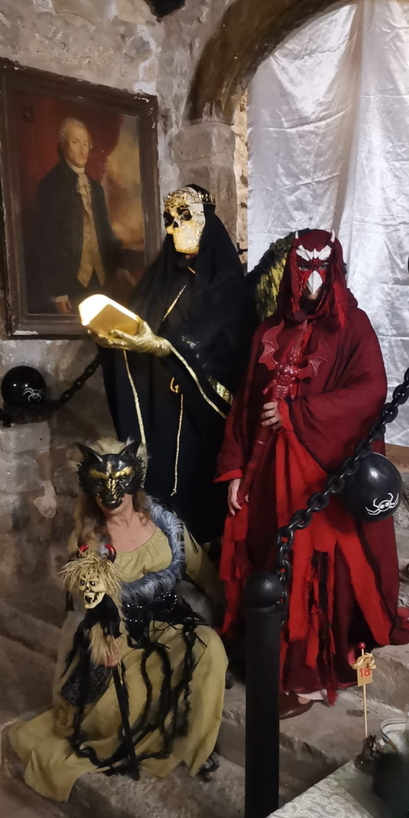 Tre personaggi in costumi evocativi per Halloween, con maschere teatrali e ambientazione storica in pietra.