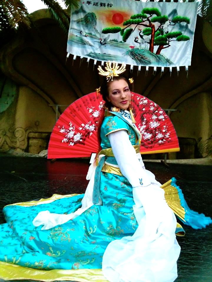 Figurante in costume orientale durante una performance esotica, con ventaglio scenico e pannello decorativo alle spalle.