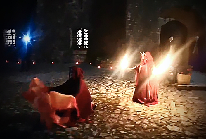 Artista in costume fantasy che danza con il fuoco in un cortile storico, con figuranti in scena.